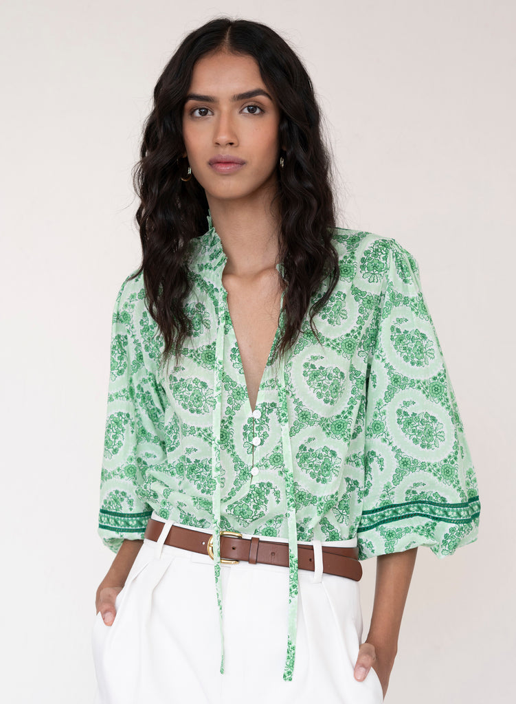 Woman wearing a green patterned blouse with white pants on a plain background