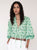 Woman wearing a green patterned blouse with white pants on a plain background